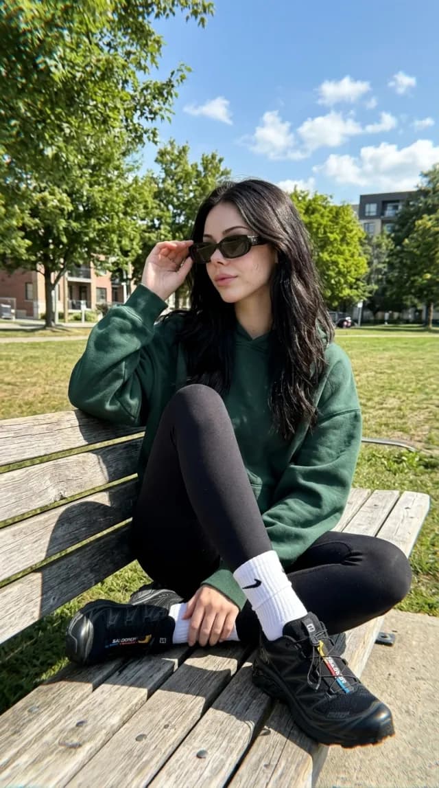 Woman in a green hoodie and black leggings sitting on a park bench in sunglasses on a sunny day